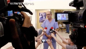 Television camras and microphones surround Dr. Ofer Merin dressed in doctor's scrubs.