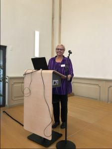 Professor Janine Geske standing at a podium with an open laptop as she addresses an audience in Germany.