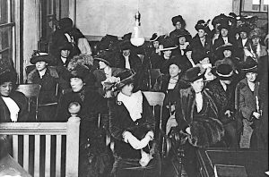 A courtroom is filled with women dressed in long black dresses and wearing hats.