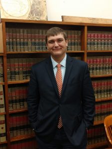 Stduent Scott Lyon, dressed in a suit, stands in front of a bookcase holding law books.