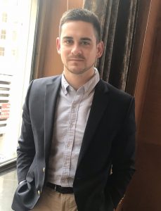 Attorney Brandon Jubelirer from the waist up stands in front of a sunlit window with his hands in his pockets