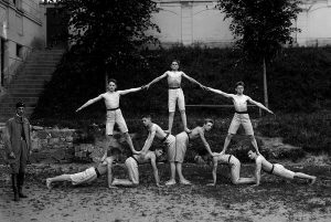 Black and white photo of a group of men in gymnast uniforms in a formation where some stand on the shoulders of others.
