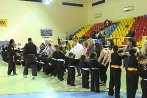 A line of young martial arts students wearing black uniforms stand with a line of adults inside of a gynmasium.