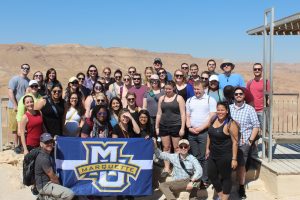 About twenty young people in casual dress surround a Marquette University flag at Masada in Israel.