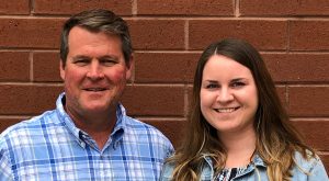 Photo of father and adult daughter standing in front of a brick wall, smiling.