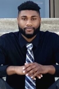 photo of a black man with short hair and a close-cropped beard wearing a black shirt with a striped tie. his hands are crossed in front of him