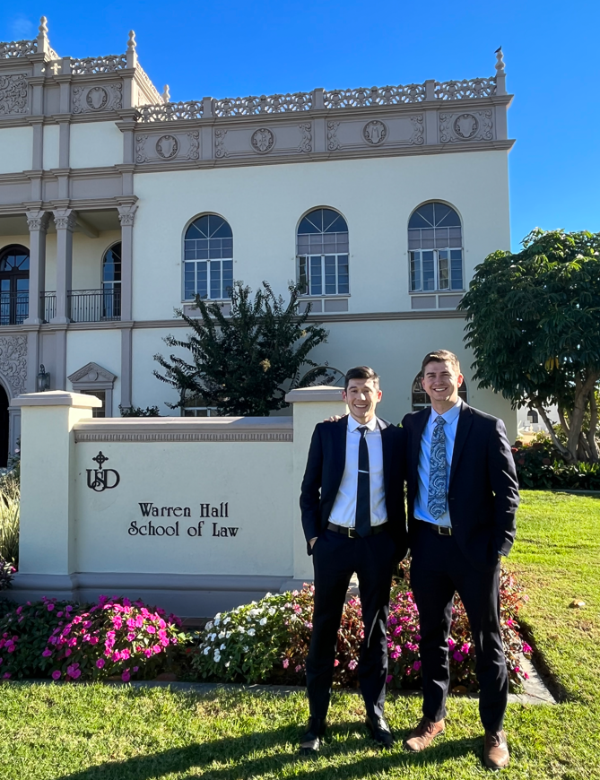 two young men in suits standing in front of the University of San Diego School of Law. One man has dark hair and the other man is taller with blonde hair. 