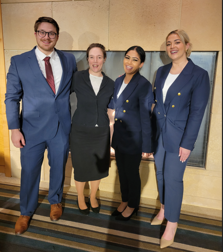 four people standing in front of a fireplace, all of them wearing business suits. On the left is a man in a blue suit and red tie. Next to him is a women in a black skirt suit, with her dark hair pulled back. Next to her is a short woman in a navy blue pantsuit with her black hair pulled back. Next to her is a blonde woman in a navy blue pantsuit. All of them are smiling big smiles.