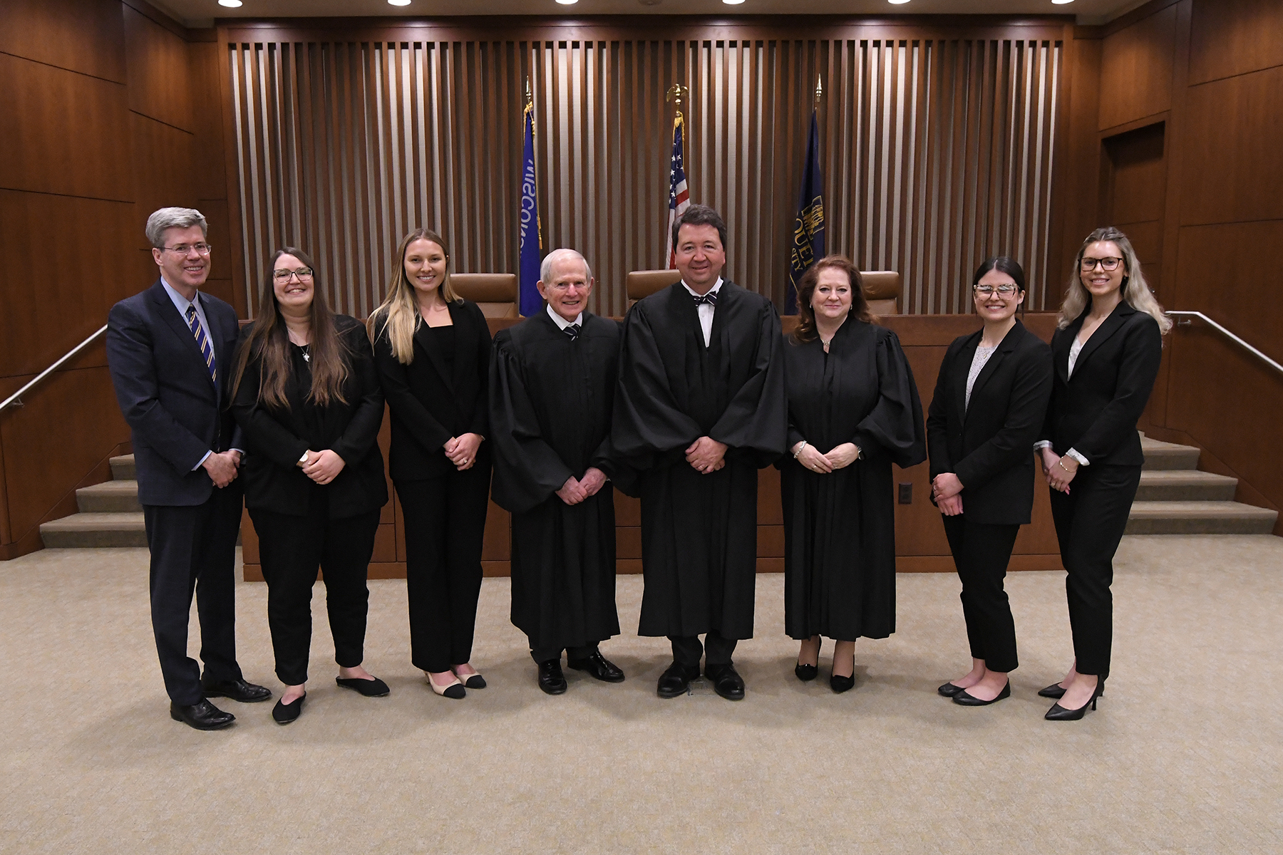8 people standing next to each other, all professionally dressed. Four are young women, three are judges, and one is the dean of the law school.